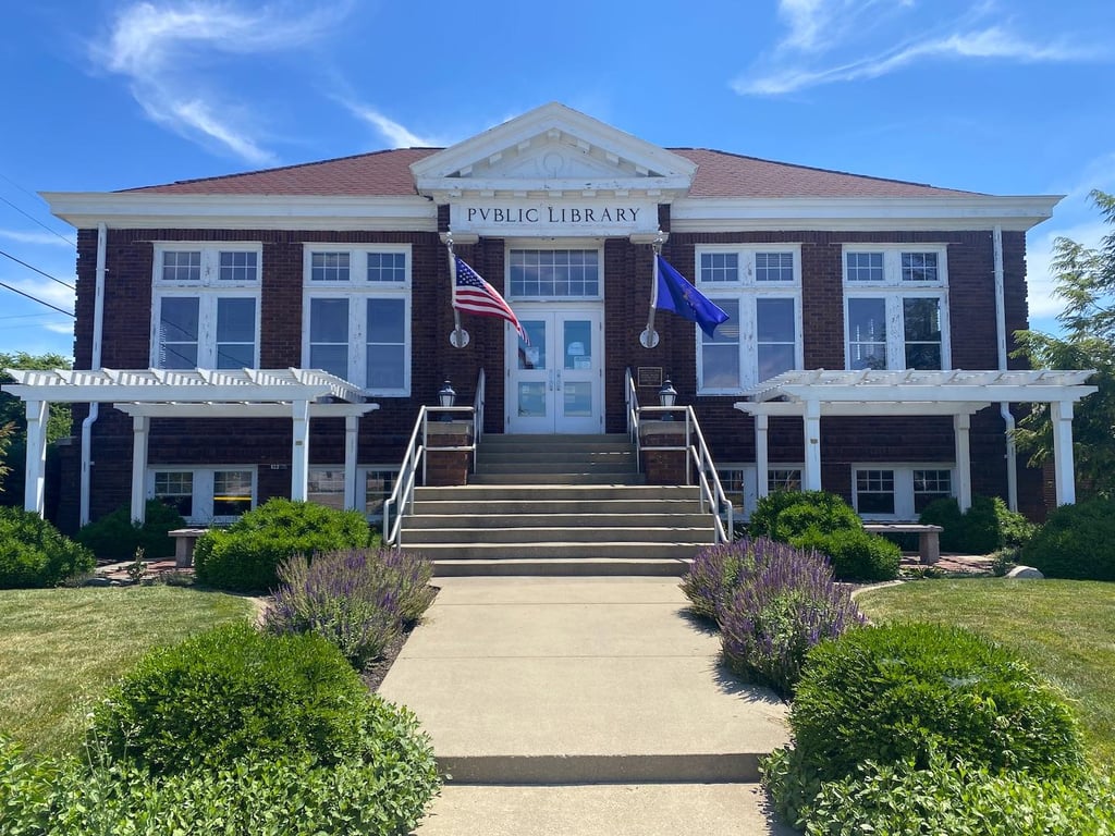 The historic Kirklin Public Library, a 1915 Carnegie-funded brick building with white columns, American and Indiana flags, and a landscaped front walkway in Kirklin, Indiana.
