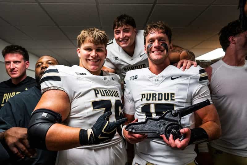 Smiling Purdue football players in white jerseys crowd the locker room as one holds the Cannon Trophy after beating Illinois in Champaign