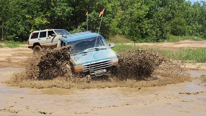 Blue SUV throwing up a wave of brown mud as it drives through a water hole, with a white Jeep Cherokee following on the trail behind it