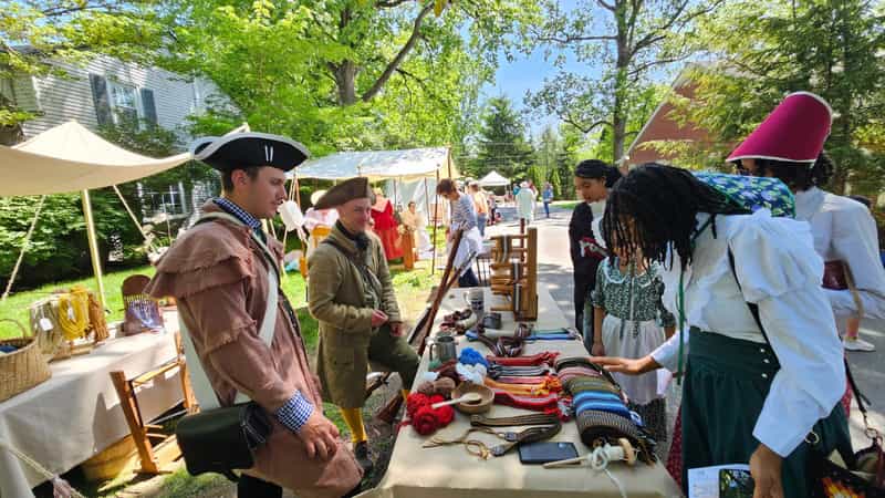 Reenactors in tricorn hats and colonial dress browse a table of handwoven sashes and yarn under shade tents at the Colonial Fair in Middletown, Indiana