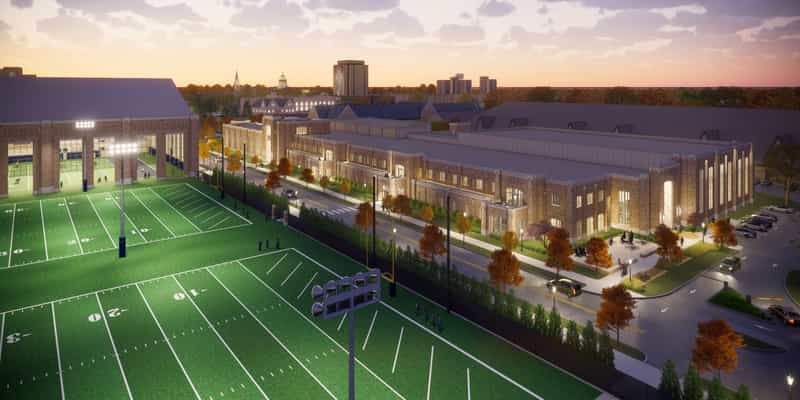 Architectural rendering at dusk of Jack and Kathy Shields Family Hall, Notre Dame's new brick football operations center, alongside the outdoor practice fields with the Golden Dome on the campus skyline behind it