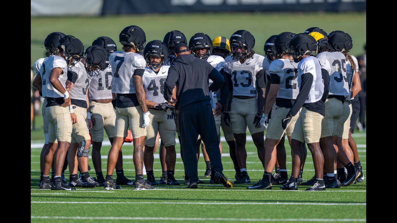 Purdue defensive players in white practice jerseys and black helmets huddled around an assistant coach on the practice field during fall camp in West Lafayette