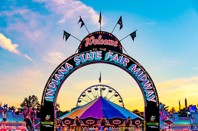 Illuminated Welcome to the Indiana State Fair Midway arch at dusk, with a Ferris wheel and carousel glowing on the midway behind it.