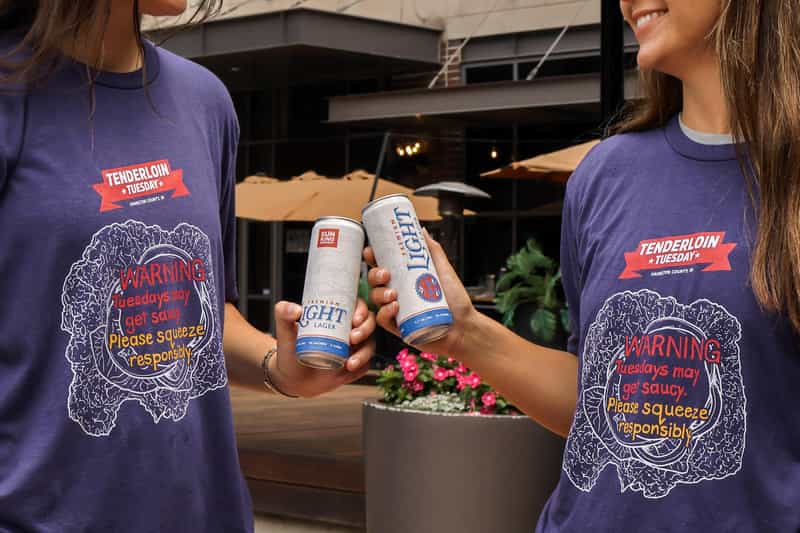Two people in purple Tenderloin Tuesday t-shirts toast cans of Sun King Premium Light Lager on a restaurant patio in Hamilton County.