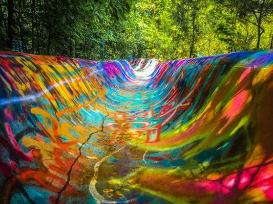 The abandoned Zoom Flume concrete waterslide near Bloomington, Indiana, its curved channel covered in vibrant multicolored graffiti with green forest behind it.