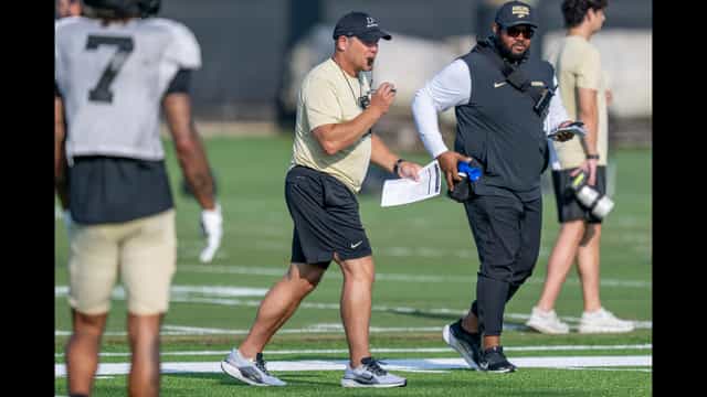 A Purdue football coach in a Boilermakers cap blows his whistle during a 2026 fall camp practice as players and staff work behind him.