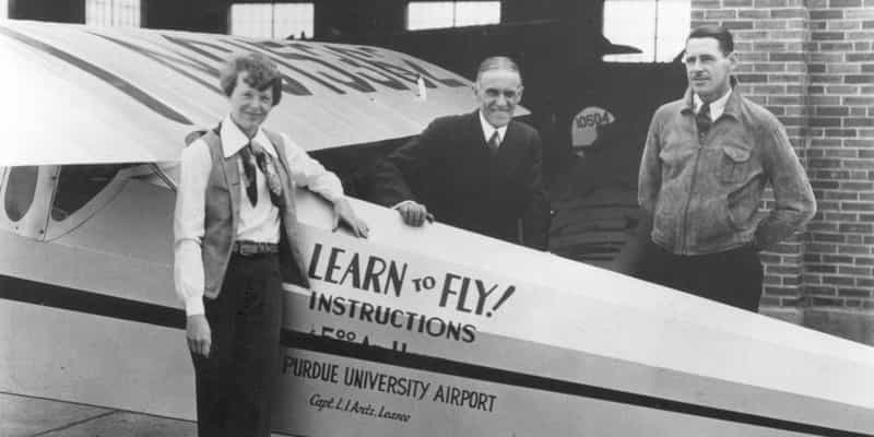 Amelia Earhart standing with two men beside a plane lettered Learn to Fly, Purdue University Airport, outside a brick hangar