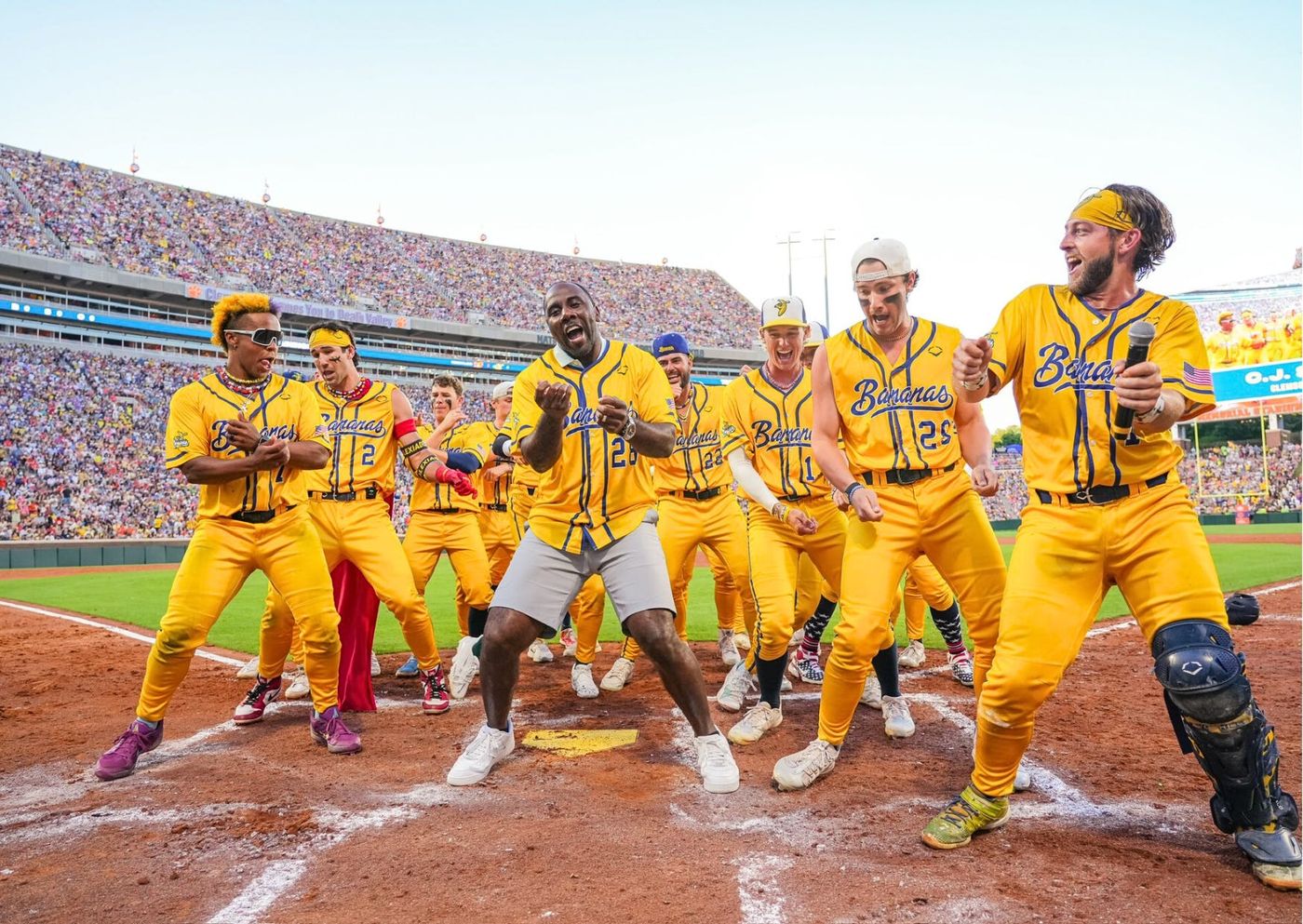 Savannah Bananas players in yellow uniforms dance around home plate during a Banana Ball game in front of a packed football stadium crowd