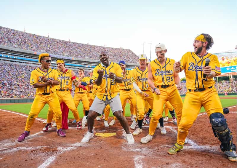 Savannah Bananas players in yellow uniforms dance around home plate during a Banana Ball game in front of a packed football stadium crowd