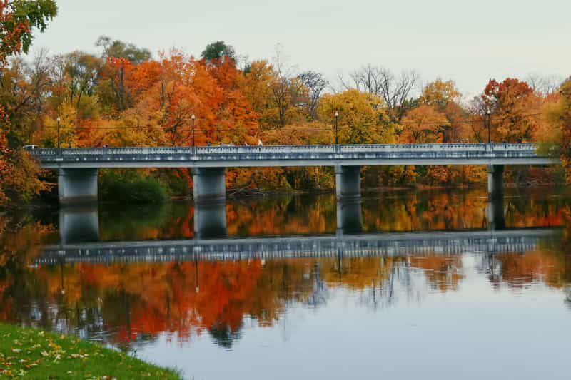 Concrete bridge over a calm river lined with orange and gold fall trees, the foliage reflected in the water