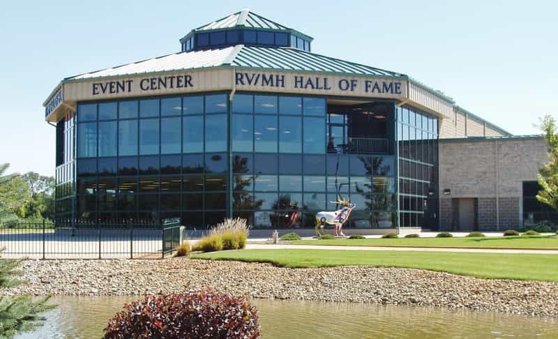 The glass-fronted exterior of the RV/MH Hall of Fame and Event Center in Elkhart, Indiana, with landscaped grounds and a pond.