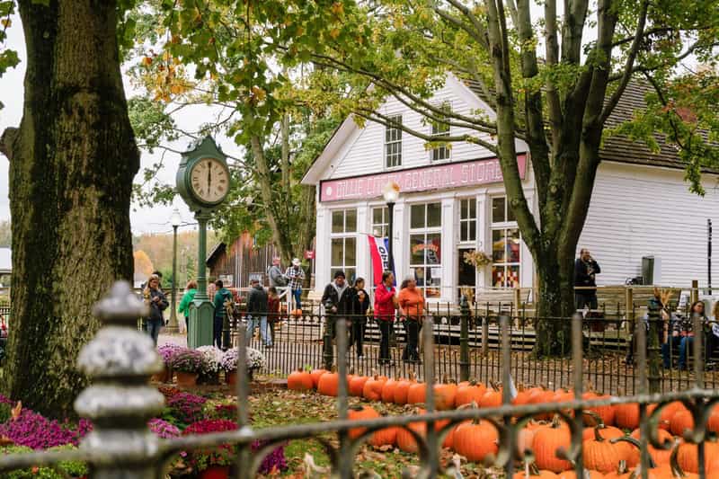 Visitors walking past the white clapboard Billie Creek General Store at Billie Creek Village near Rockville, Indiana, with a green street clock, mums and rows of pumpkins in the foreground on a fall day