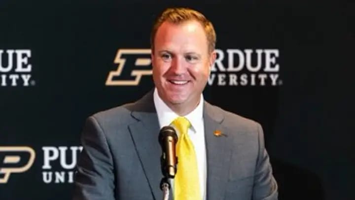 Tommy McClelland smiles at a podium during his introduction as Purdue's vice president and director of intercollegiate athletics, in front of a Purdue University backdrop.
