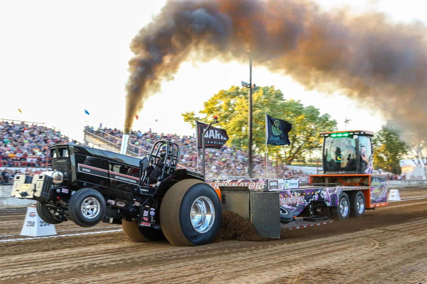 A modified pull tractor lifts its front wheels and throws diesel smoke as it drags a weight-transfer sled down a dirt track before a packed grandstand at an Indiana county fair.