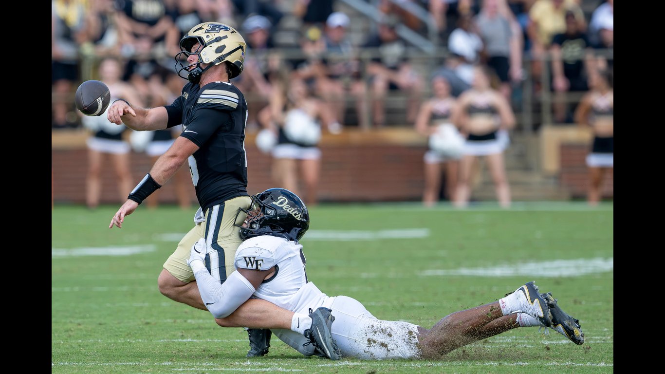 A Purdue quarterback in a gold helmet releases a pass as a Wake Forest defender dives to tackle him at Ross-Ade Stadium