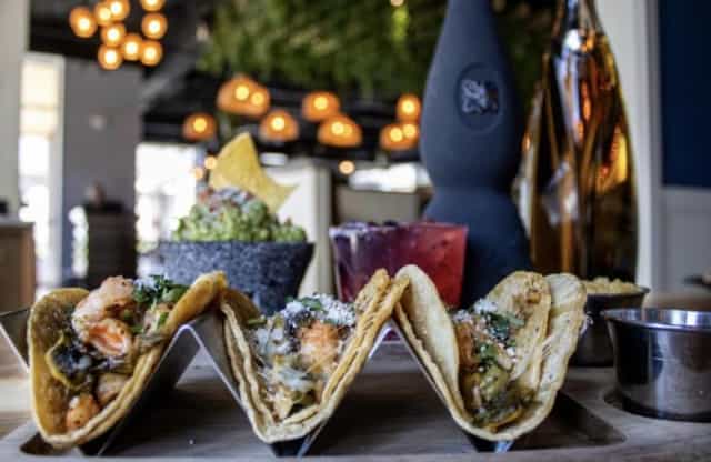Three street tacos topped with shrimp, cilantro and cotija on a metal rack at Tlahcos Mexican Cuisine in Plainfield, Indiana, with guacamole and a red cocktail behind them