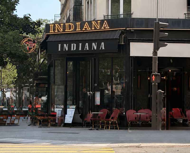 A Paris corner restaurant with a glowing red INDIANA sign and neon logo above a black awning, with empty red bistro chairs out front on a bright summer day