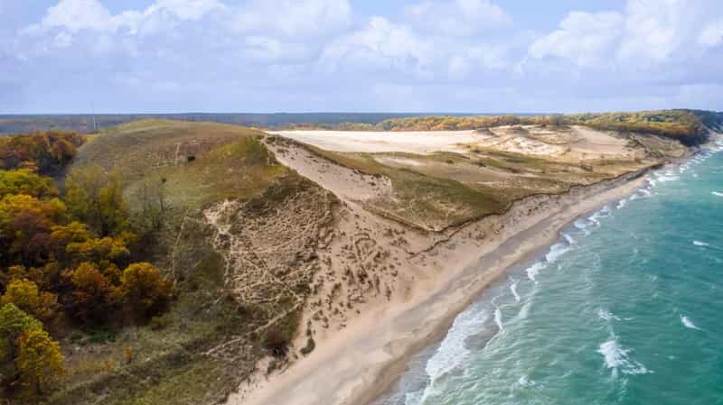 Aerial view of a tall sand dune and fall-colored woods above a beach along the Lake Michigan shoreline, with waves rolling in