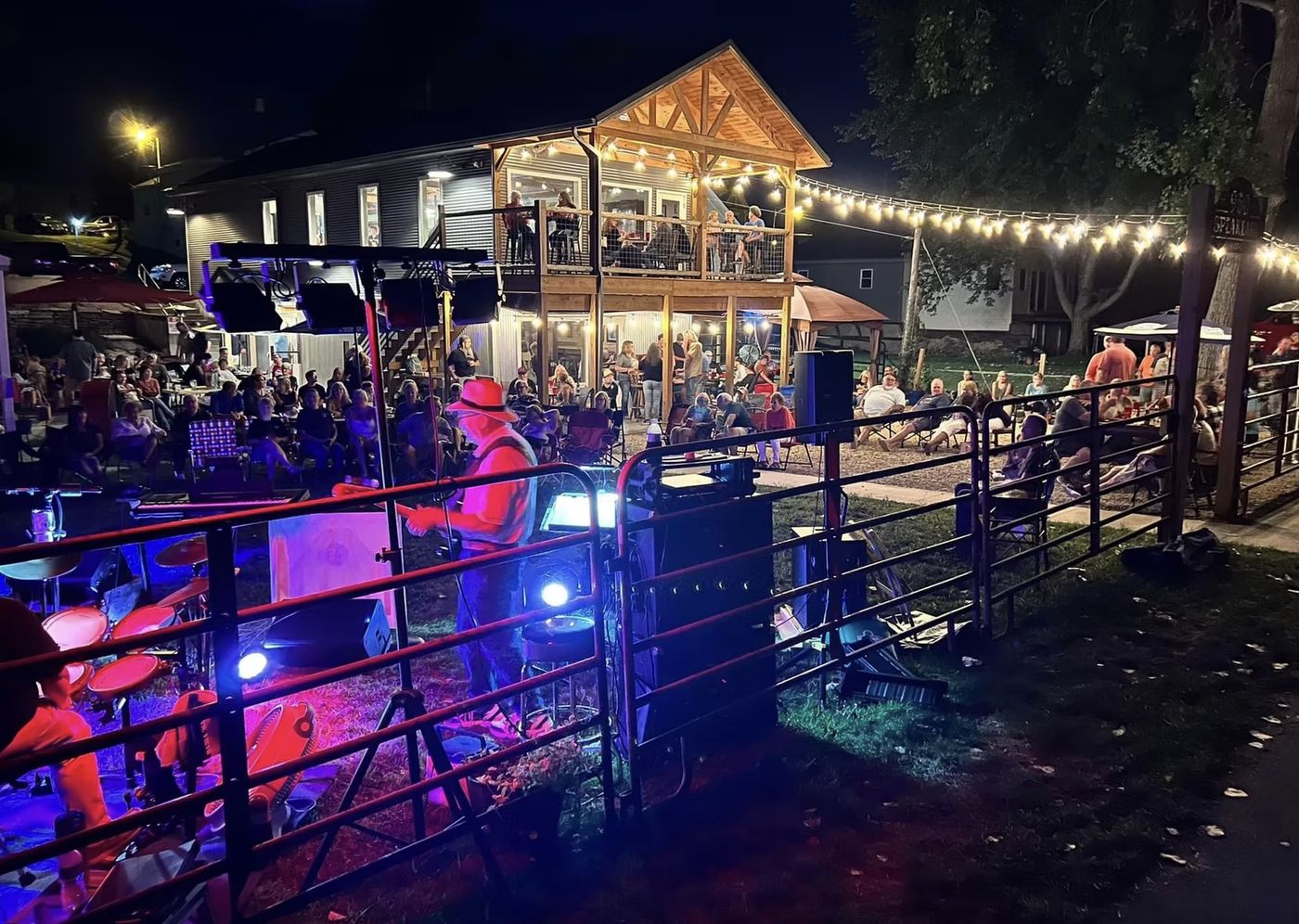 Nighttime crowd gathered on the lawn at 950 Speakeasy Bistro in Lagro, Indiana, watching a live band play under string lights beside the two-story timber patio