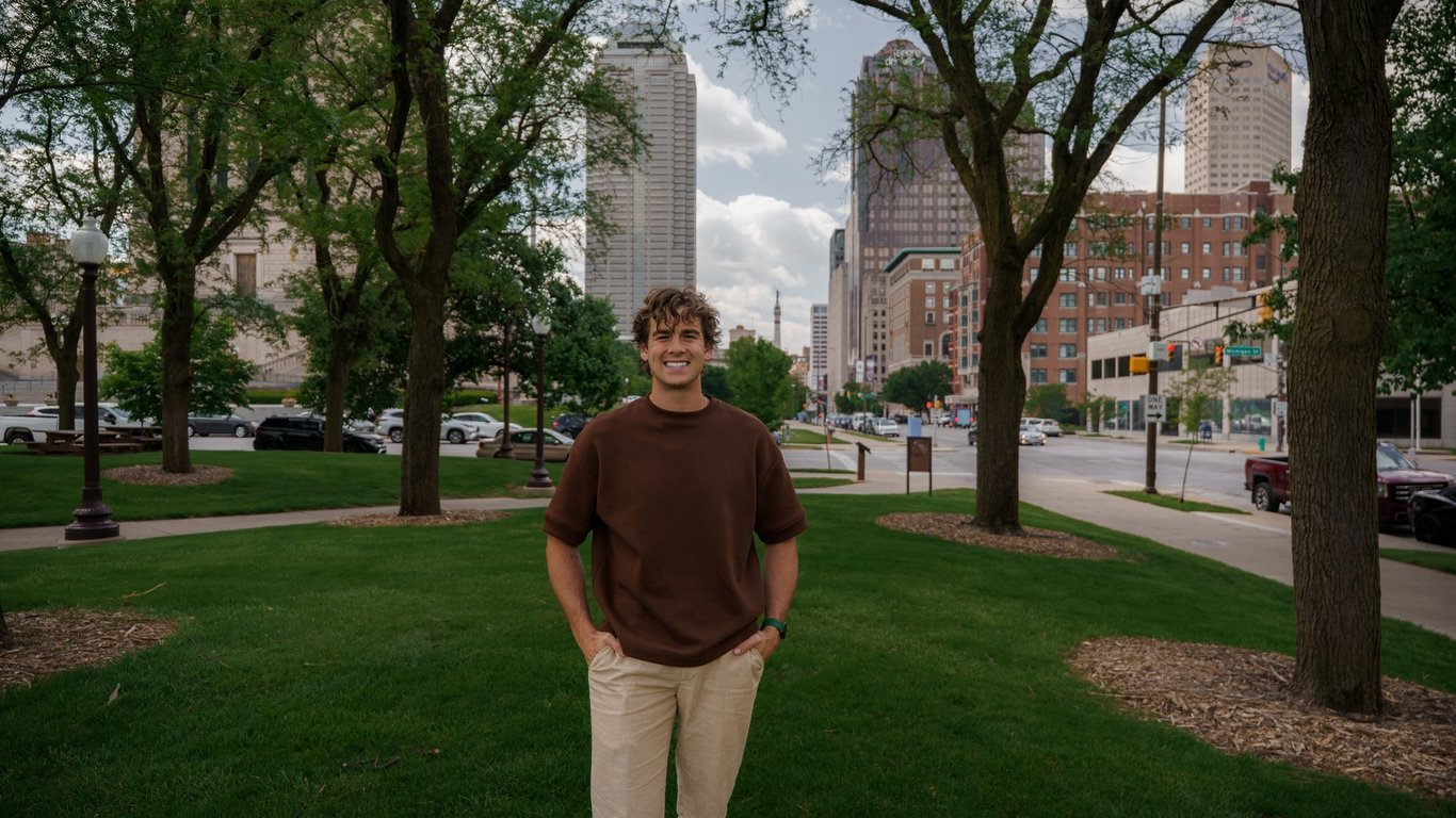 Ian DeFelice, a Roots Realty Co. agent at @properties who works with first-time buyers, standing in a downtown Indianapolis park with city buildings behind him