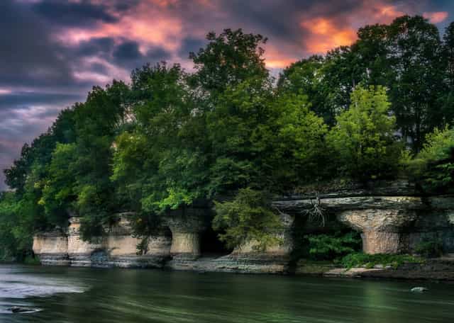 Limestone pillars and shallow alcoves carved into a wooded bluff above the Mississinewa River near Peru, Indiana, under a pink and orange sunset sky