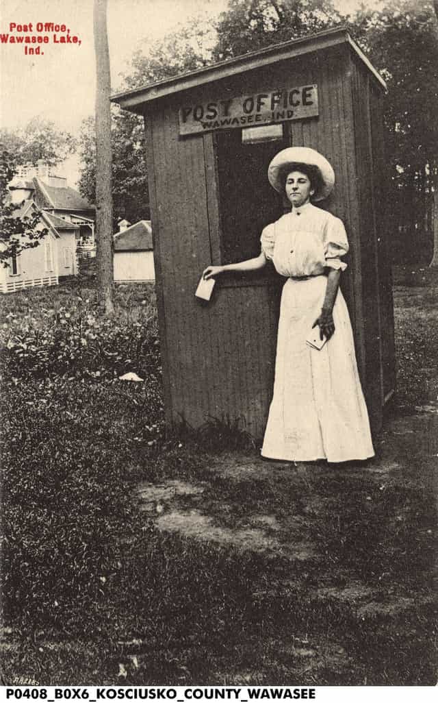 Vintage postcard of a woman in a white dress and wide hat mailing a letter at the tiny wooden Wawasee, Ind. post office