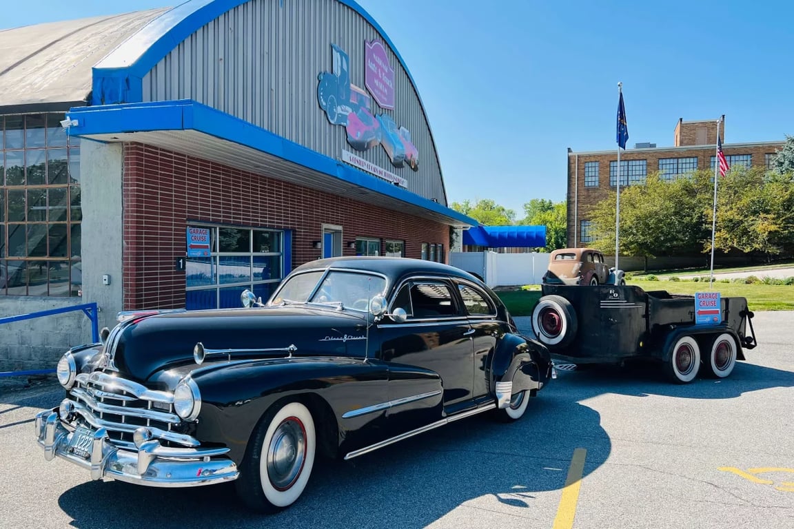 Black vintage coupe and weathered custom truck parked outside an Auburn automotive venue during the Annual Garage Cruise