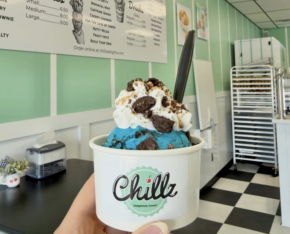 A hand holding a Chillz Delights cup of blue ice cream topped with whipped cream and brownie chunks inside the shop's mint-green interior in North Manchester, Indiana