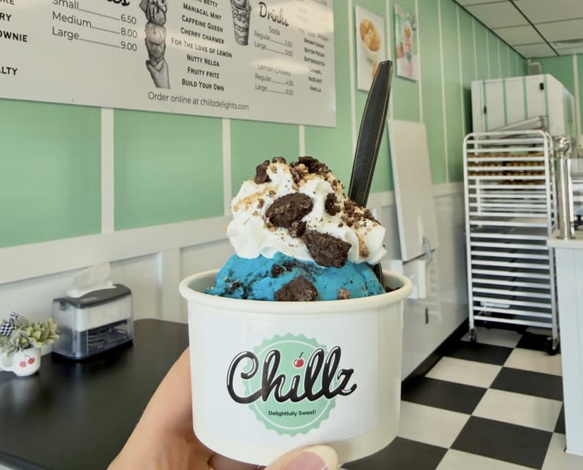 A hand holding a Chillz Delights cup of blue ice cream topped with whipped cream and brownie chunks inside the shop's mint-green interior in North Manchester, Indiana