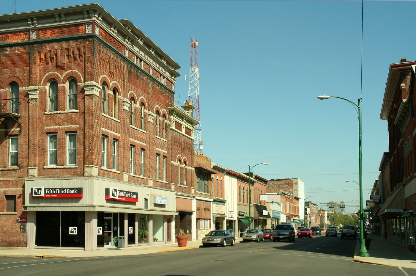 Historic brick storefronts along a downtown street in Decatur, Indiana, with a three-story corner building and US 224 route signs at the intersection ahead