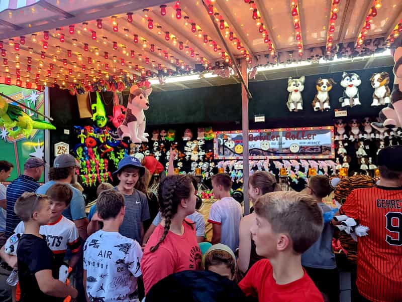 Kids crowd a carnival game booth at the Bluffton Free Street Fair at night, under rows of red string lights, with stuffed huskies and other prizes hanging above a Bowler Roller game.