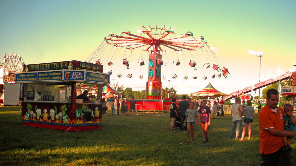 Fairgoers ride a swing carnival ride at dusk beside a Fresh Fried Veggies food stand at an Indiana county fair.