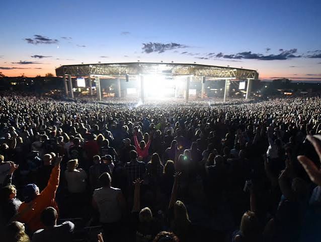 A packed amphitheater crowd faces bright stage lights at dusk during an outdoor summer concert in Indiana, hands raised under a pink and blue sky.