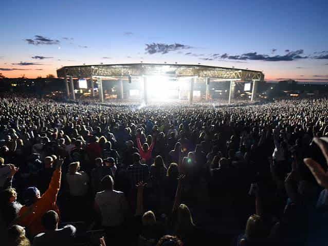 A packed amphitheater crowd faces bright stage lights at dusk during an outdoor summer concert in Indiana, hands raised under a pink and blue sky.