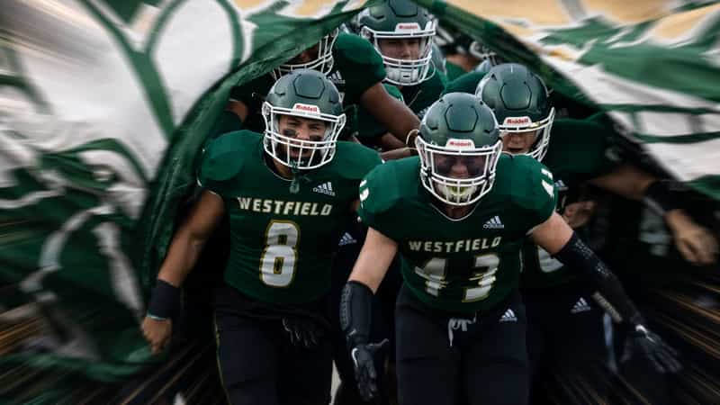 Westfield Shamrocks football players in green jerseys and helmets run through a green and white banner onto the field before a game