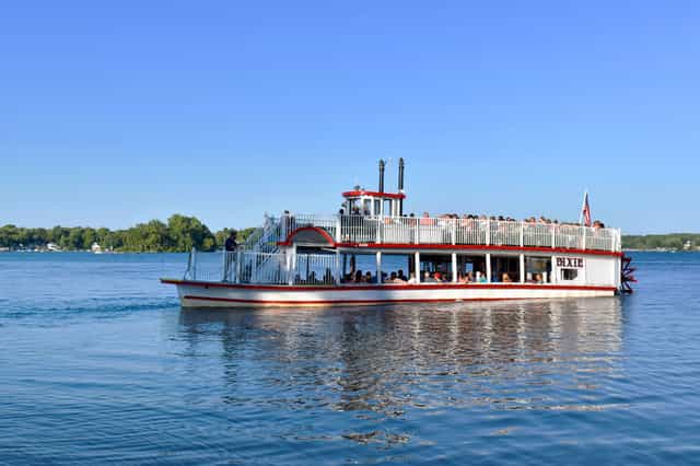 The Dixie, a red-and-white sternwheel paddleboat, cruising across Webster Lake in North Webster, Indiana, with passengers on its decks under a clear blue sky.