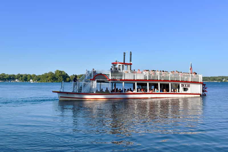 The Dixie, a red-and-white sternwheel paddleboat, cruising across Webster Lake in North Webster, Indiana, with passengers on its decks under a clear blue sky.