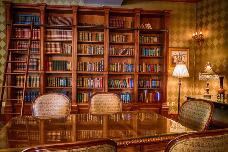 Wood-paneled library with floor-to-ceiling bookshelves, a rolling ladder, table lamps and a glass-topped reading table