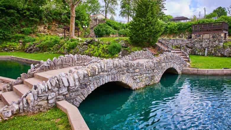 Stone arch footbridge crossing the turquoise water at the Sunken Gardens in Huntington, Indiana, with limestone stairways, green lawns, and a wooden overlook deck above the former quarry walls
