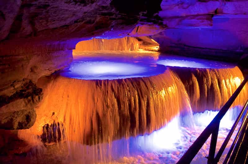 An illuminated rimstone formation at Squire Boone Caverns, its terraced flowing stone lit in blue and amber as water cascades over the ledges.