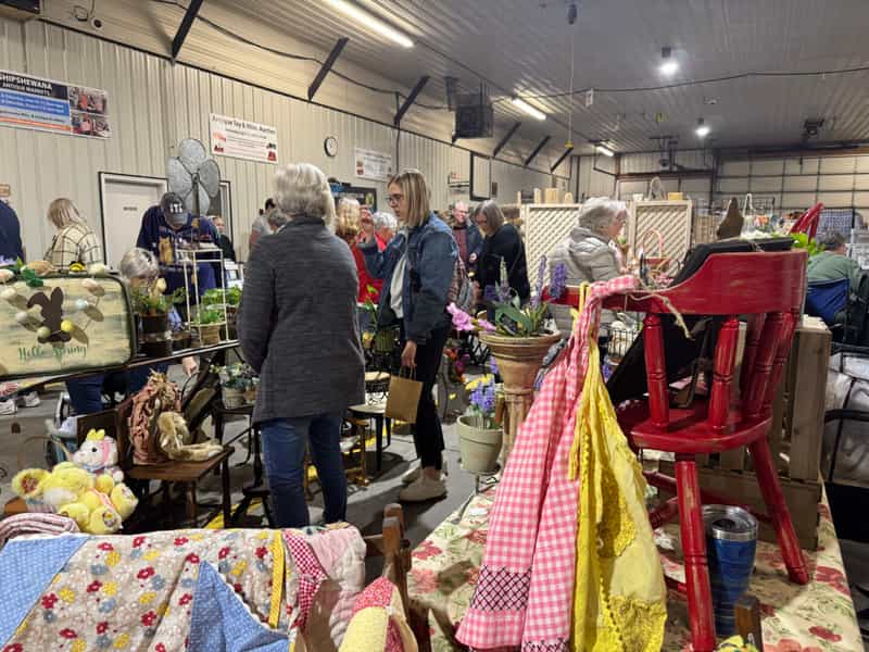 Shoppers browsing crowded craft and vintage booths (quilts, a red wooden chair, home decor) inside the auction building