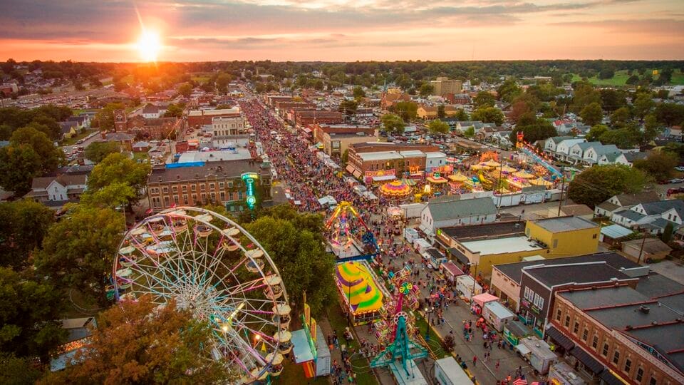 Aerial sunset view of the West Side Nut Club Fall Festival, with a Ferris wheel and carnival rides lining a crowded Franklin Street in Evansville