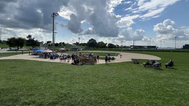 Players and spectators on the original Field of Dreams baseball diamond in Dyersville, Iowa, with the new permanent stadium visible beyond the outfield corn.