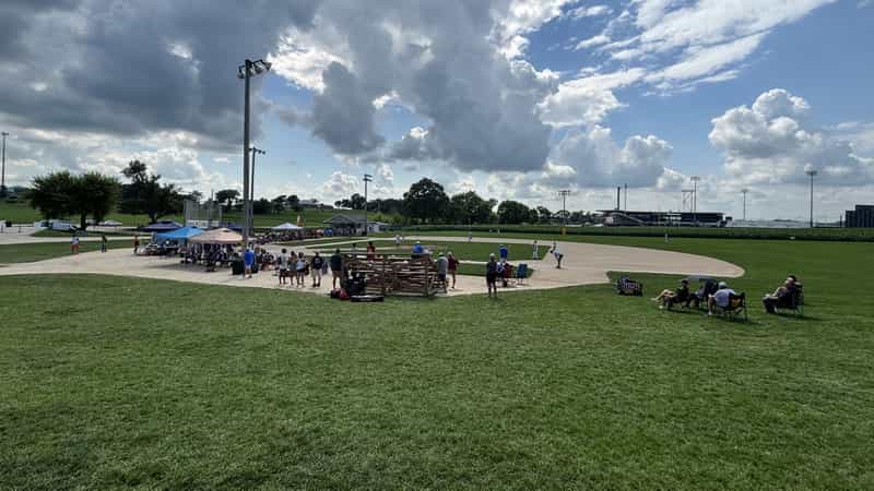 Players and spectators on the original Field of Dreams baseball diamond in Dyersville, Iowa, with the new permanent stadium visible beyond the outfield corn.