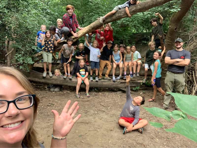 Children and instructors gather on a fallen log in the woods during a White Pine Wilderness Academy forest school session in Indianapolis.
