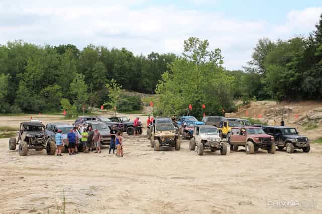 Lifted Jeeps, a Cherokee and a rock crawler lined up on an open sand flat beside wooded hills while riders gather between the vehicles