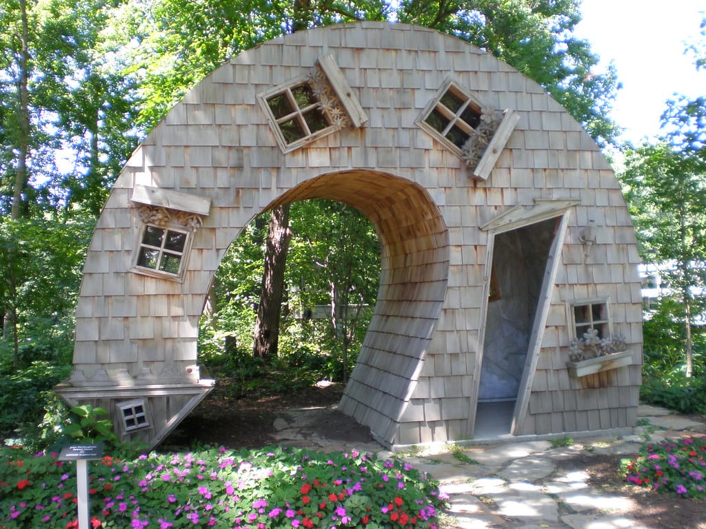 The Twisted House sculpture at the Indianapolis Art Center, a cedar-shingled house bent into an arch with tilted windows and a crooked open doorway, behind a bed of pink and red flowers