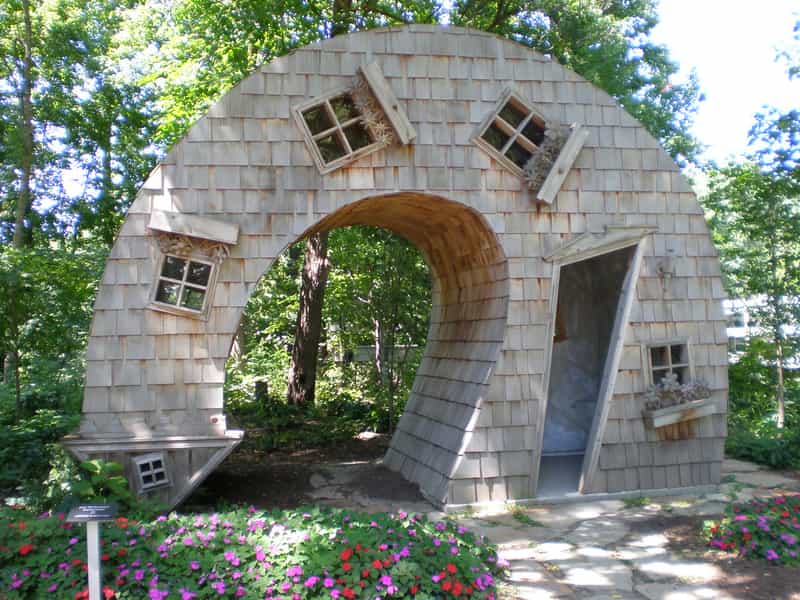 The Twisted House sculpture at the Indianapolis Art Center, a cedar-shingled house bent into an arch with tilted windows and a crooked open doorway, behind a bed of pink and red flowers