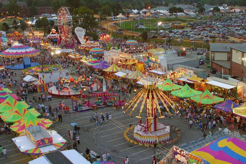 Aerial view of a packed Indiana fairground at dusk, filled with a Ferris wheel, swing rides, and colorful midway tents.