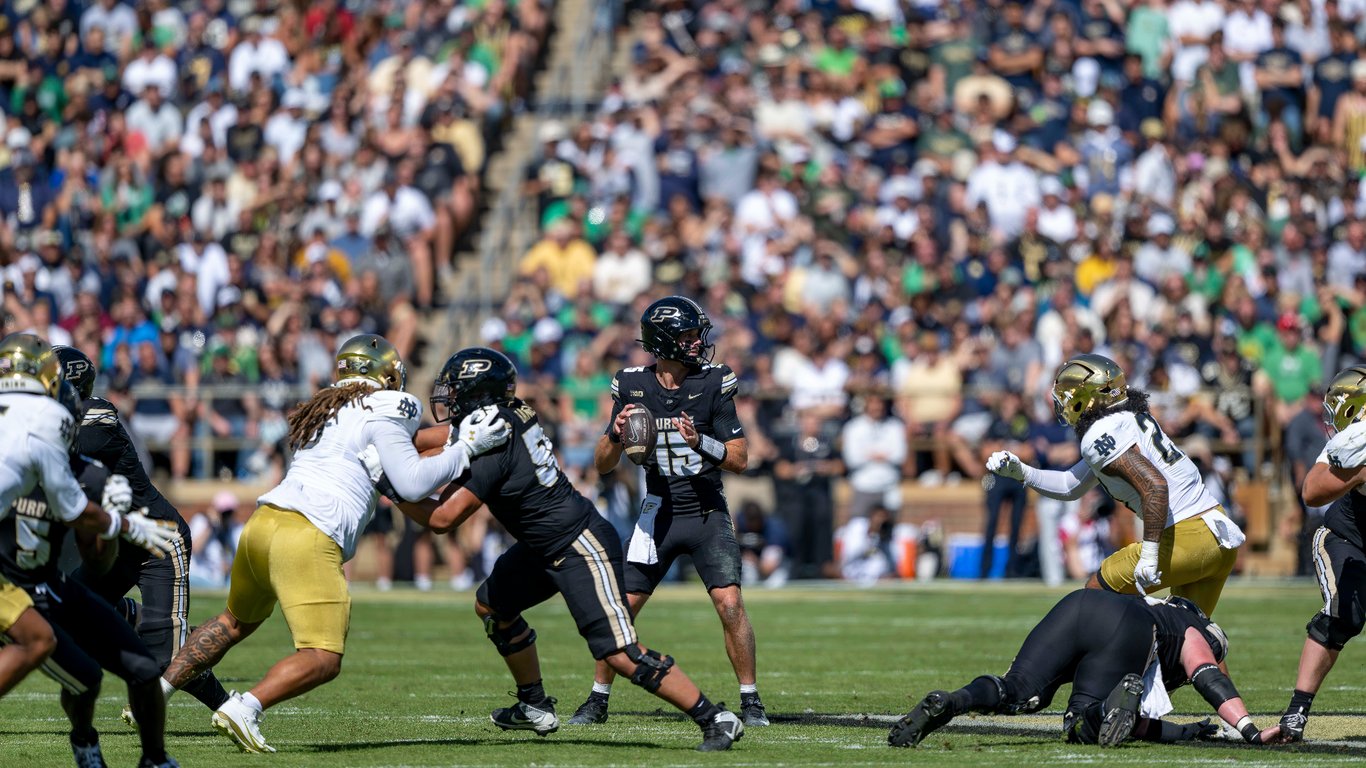 Purdue quarterback wearing No. 15 drops back to pass as Notre Dame defenders rush in front of a packed Ross-Ade Stadium crowd